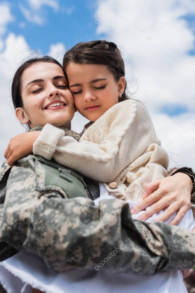 smiling military woman with closed eyes embracing daughter against cloudy sky