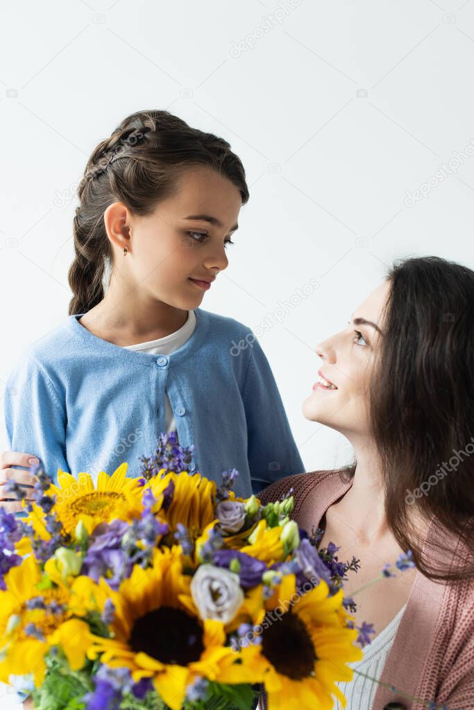 mom and daughter smiling at each other near blue and yellow flowers isolated on grey