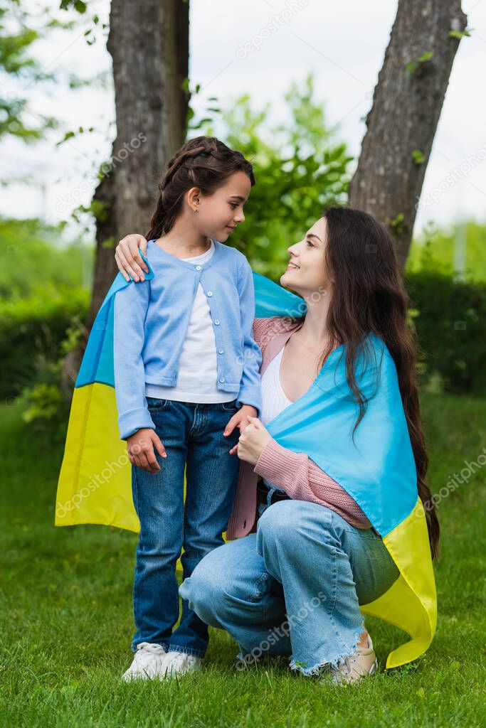 mom and daughter covered with ukrainian flag smiling at each other in park