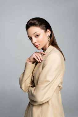 brunette young model in beige blazer looking at camera isolated on grey