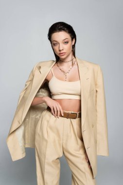  young woman in stylish beige outfit looking at camera while posing isolated on grey
