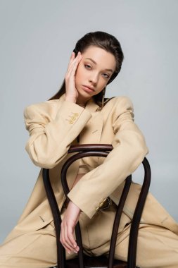 young woman in stylish outfit looking at camera while sitting on chair isolated on grey