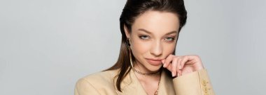 sly young woman in stylish blazer looking at camera while posing isolated on grey, banner