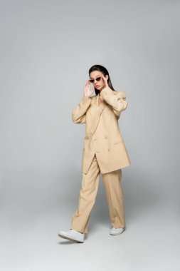 full length of young woman in stylish beige outfit wearing trendy sunglasses and walking on grey