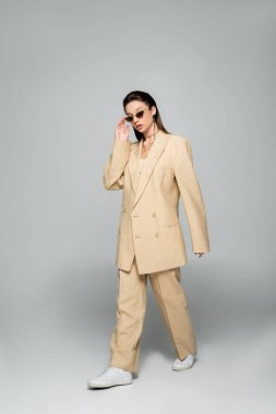 full length of young woman in stylish beige suit wearing trendy sunglasses and walking on grey