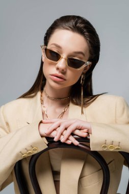 young woman in stylish outfit and trendy sunglasses sitting on wooden chair isolated on grey