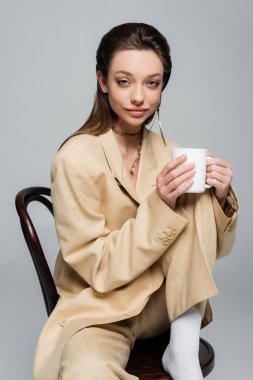 brunette woman in stylish beige suit holding cup of coffee and sitting on wooden chair isolated on grey