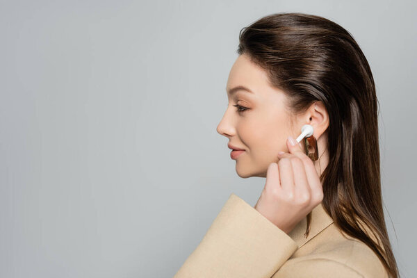 side view of happy young woman in stylish outfit wearing wireless earphone isolated on grey
