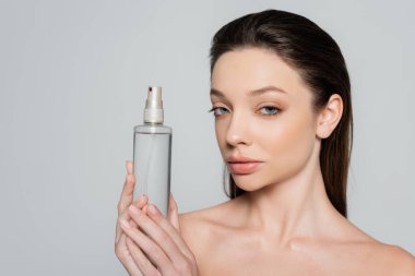 brunette young woman with bare shoulders holding bottle with cosmetic product isolated on grey