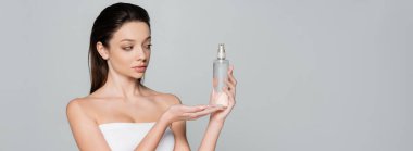 young woman with bare shoulders holding bottle with cosmetic product isolated on grey, banner