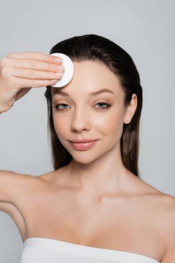 happy young woman removing makeup on forehead with cotton pad isolated on grey