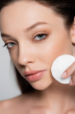 close up view of young woman removing makeup with cotton pad isolated on grey