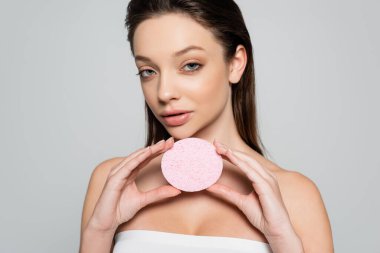 brunette young woman with bare shoulders holding exfoliating sponge isolated on grey