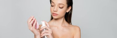 young woman with bare shoulders holding bottle with cosmetic product near exfoliating sponge isolated on grey, banner