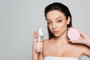 young woman holding bottle with cosmetic product near exfoliating sponge isolated on grey