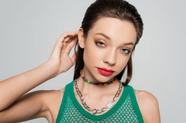 young and stylish woman with red lips adjusting hair while posing isolated on grey