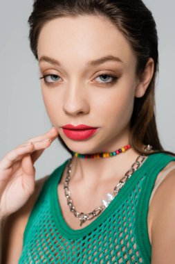 close up view of young and stylish woman with red lips touching necklace while posing isolated on grey