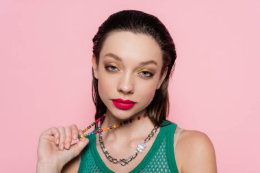 young brunette woman with bright makeup pulling beads necklace while looking at camera isolated on pink 