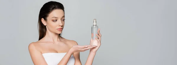 young woman with bare shoulders holding bottle with cosmetic product isolated on grey, banner