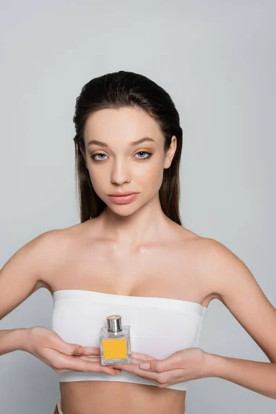 young woman with bare shoulders holding bottle with perfume isolated on grey