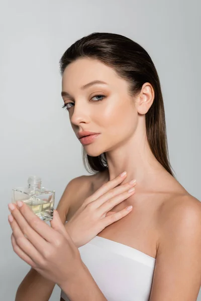 brunette woman with naked shoulders holding bottle with luxury perfume isolated on grey