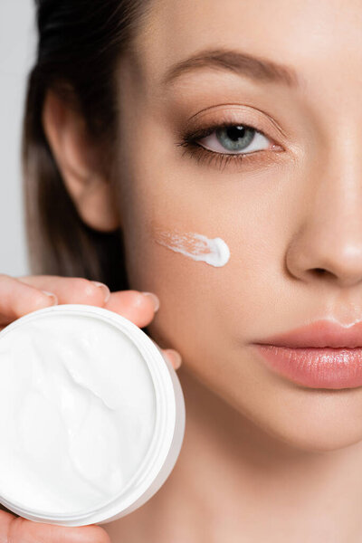 close up view of cropped young woman with cosmetic cream on cheek holding container isolated on grey