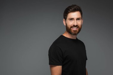 portrait of smiling bearded man in black t-shirt looking at camera isolated on grey