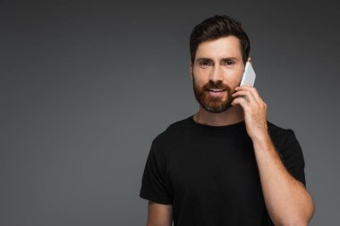 cheerful and bearded man in black t-shirt talking on smartphone isolated on grey