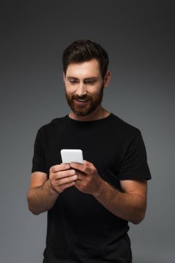 happy bearded man in black t-shirt messaging on smartphone isolated on grey