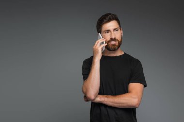 positive and bearded man in black t-shirt talking on smartphone isolated on grey