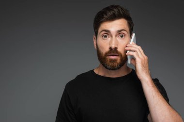 shocked man in black t-shirt talking on smartphone isolated on grey