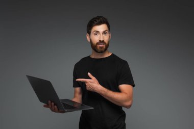 amazed and bearded man in black t-shirt pointing with finger at laptop isolated on grey