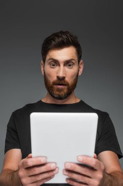 shocked and bearded man in black t-shirt holding digital tablet isolated on grey