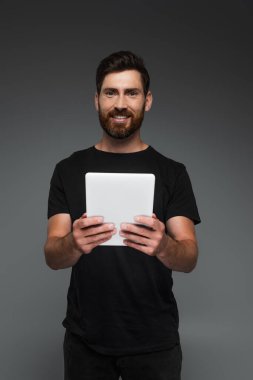cheerful and bearded man in black t-shirt holding digital tablet isolated on grey
