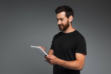 cheerful bearded man in black t-shirt using digital tablet isolated on grey