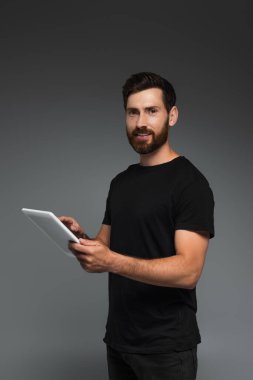 happy and bearded man in black t-shirt holding digital tablet isolated on grey