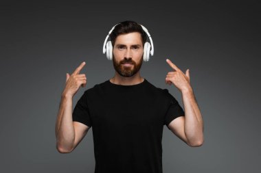 bearded man listening music and pointing with fingers at wireless headphones isolated on grey