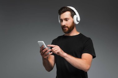 bearded man in wireless headphones listening music and pointing at smartphone isolated on grey