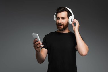 smiling man in wireless headphones listening music and using smartphone isolated on grey