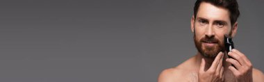 cheerful and shirtless man with hair on chest shaving beard with electric razor isolated on grey, banner
