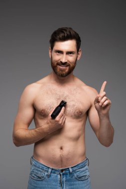 happy and shirtless man shaving hair on chest with electric razor and gesturing isolated on grey