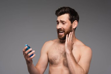 bearded and shirtless man holding bottle with after shave product isolated on grey