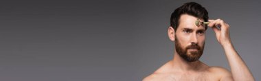 shirtless man with beard using jade roller while massaging face isolated on grey, banner