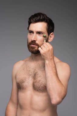 shirtless man with beard using jade roller while massaging face isolated on grey