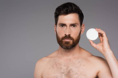 bearded and shirtless man holding container with cosmetic cream isolated on grey