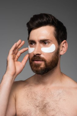 bearded man applying moisturizing eye patches isolated on grey 