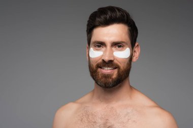 happy bearded man with moisturizing eye patches isolated on grey 