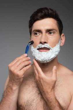 shirtless man with white shaving foam on face shaving with safety razor on grey clipart
