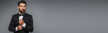 bearded man in black tuxedo with bow tie adjusting sleeve on white shirt isolated on grey, banner