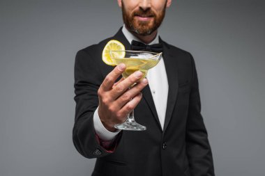 cropped view of cheerful and bearded man in suit with bow tie holding glass of cocktail isolated on grey